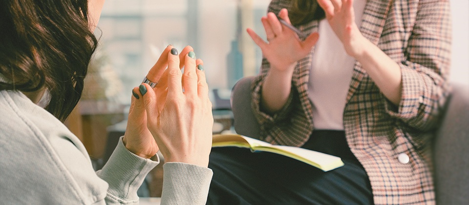 one woman with a notepad explains something to a woman who presses her fingers together to illustrate the concept of a consultant giving input to an engineer