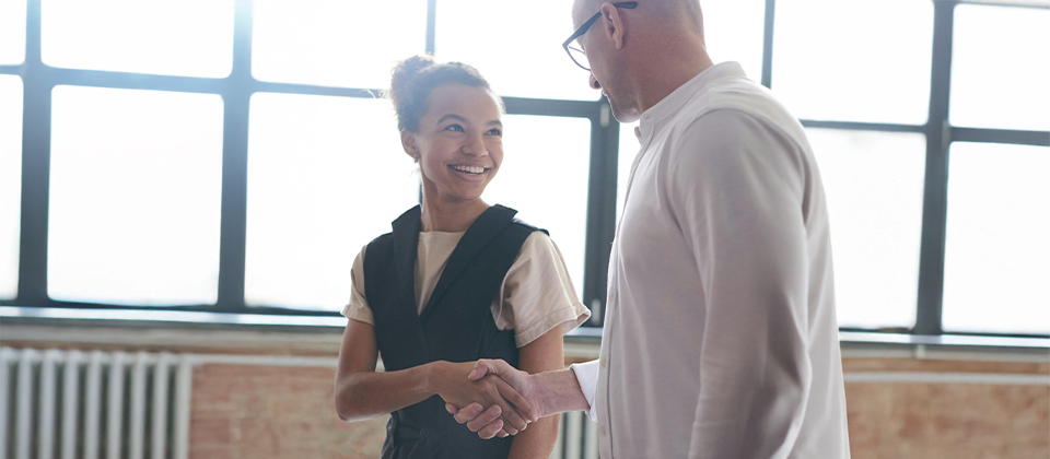 An older businessman shakes the hand of a younger businesswoman to illustrate the concept of succession planning